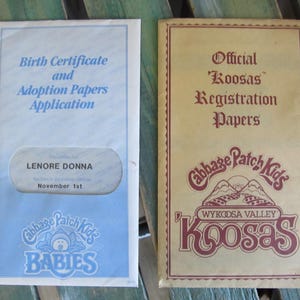May include: Two Cabbage Patch Kids registration papers. The left paper is blue and white with the text "Birth Certificate and Adoption Papers Application" and "LENORE DONNA November 1st". The right paper is brown with the text "Official Koosas Registration Papers" and "Cabbage Patch Kids WYKODSA VALLEY Koosas".
