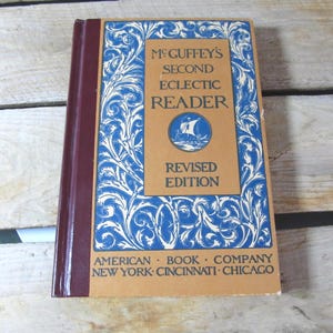 May include: A vintage book titled "McGuffey's Second Eclectic Reader, Revised Edition." The cover features a tan central panel with blue text and a sailboat illustration, framed by a blue and white ornate design. The spine is a deep burgundy color.