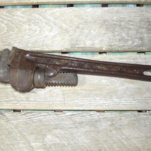 Op de afbeelding: Een vintage, roestbruine pijpsleutel, liggend op een verweerd houten oppervlak. De sleutel heeft een verstelbare bek en een lange handgreep met een gat aan het uiteinde. De tool lijkt gebruikt en vertoont tekenen van ouderdom.