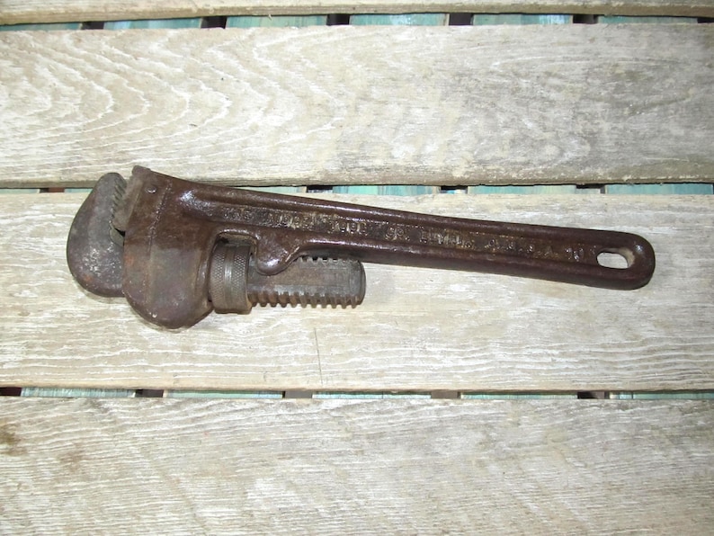 Op de afbeelding: Een vintage, roestbruine pijpsleutel, liggend op een verweerd houten oppervlak. De sleutel heeft een verstelbare bek en een lange handgreep met een gat aan het uiteinde. De tool lijkt gebruikt en vertoont tekenen van ouderdom.