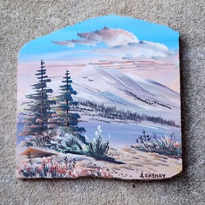 May include: A hand-painted landscape painting on a wooden plaque depicting a snowy mountain scene with two pine trees in the foreground. The sky is blue with a pink and white cloud. The painting is signed "A.O'Henry".