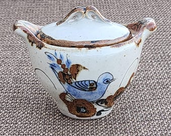 May include: A ceramic lidded bowl with a hand-painted design. The bowl is off-white with a blue bird and brown floral accents. The lid has a handle and a brown rim. The bowl is likely handmade and has a rustic aesthetic.