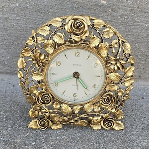 May include: A gold-toned metal alarm clock featuring an intricate filigree frame decorated with sculpted roses and leaves. The round clock face is off-white with gold numerals, the text MATSON and ALARM, and luminous hands indicating the time.