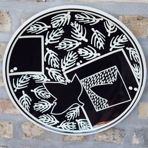 May include: A black and white ceramic plate with a geometric design. The plate features a stylized leaf shape and a pattern of small branches.