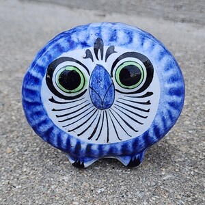 May include: A ceramic owl figurine with a blue and white design. The owl has large, round eyes with black pupils and green irises. Black lines radiate from the eyes, and the owl has small black feet. The figurine is a decorative item.