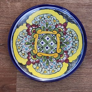 May include: A round, hand-painted ceramic plate with a vibrant floral design. The plate features a dark blue rim, a yellow central design, and a detailed pattern of flowers in green, red, and white. The plate is approximately 10 inches in diameter.