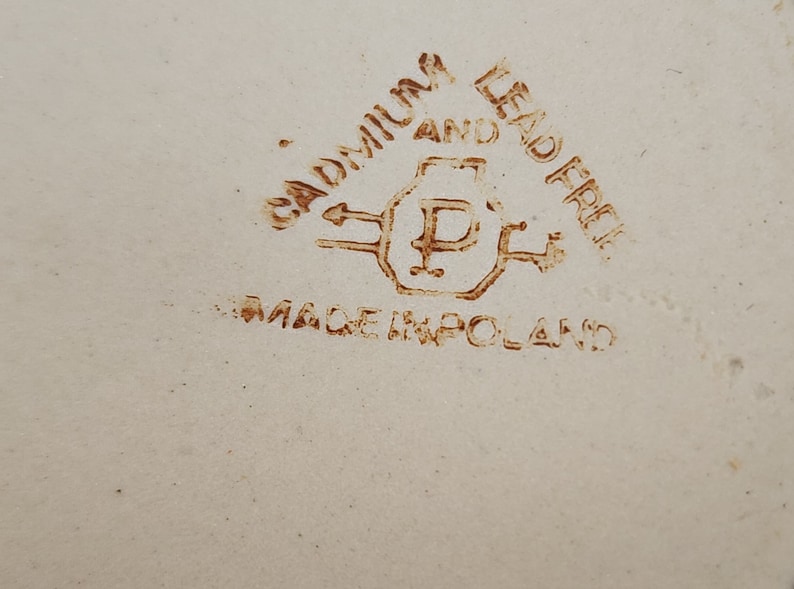 Op de afbeelding: Close-up van een bruine stempel op een wit oppervlak. De stempel luidt "Cadmium and Lead Free" met een pijl die naar links wijst en een pijl die naar rechts wijst. In het midden van de stempel staat een letter "P" in een vierkant. Onder de stempel staat de tekst "Made in Poland".