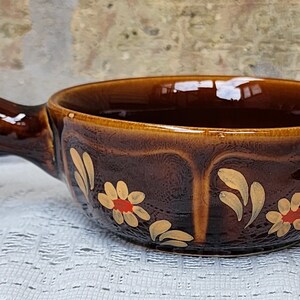 Op de afbeelding: Een bruine keramische soepkom met een handvat. De kom is versierd met een bloemmotief in goud en rood.