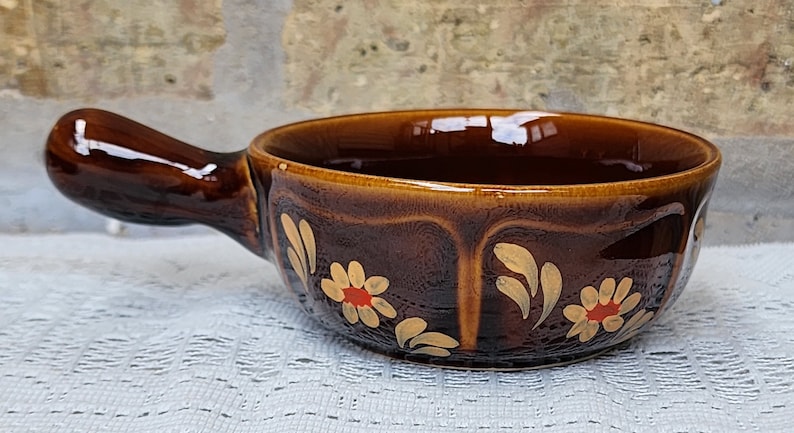 Op de afbeelding: Een bruine keramische soepkom met een handvat. De kom is versierd met een bloemmotief in goud en rood.