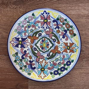 May include: A white ceramic plate with a blue, yellow, green, and pink floral design. The plate has a scalloped edge and is decorated with a repeating pattern of flowers and leaves.
