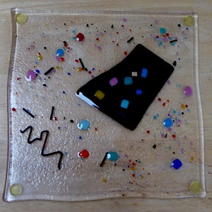 May include: A square glass coaster with a black geometric shape in the center. The coaster is decorated with colorful dots and lines.