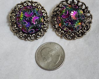 May include: A pair of vintage clip-on earrings with iridescent centres and ornate silver-tone filigree frames. A quarter coin is included for size comparison. The earrings have a floral pattern. The earrings are approximately 2.5 cm in diameter.