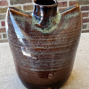 May include: A brown ceramic vase with a unique, asymmetrical shape and a light green glaze. The vase has a wide opening and a narrow base.