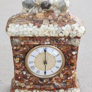 May include: A decorative clock with a stone-like exterior. The clock face has Roman numerals and gold-colored hands. The clock is made of various small stones in shades of brown, beige, and white. The clock is a rectangular shape with a curved top.
