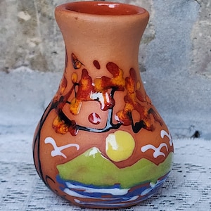 May include: A small, hand-painted terracotta vase with a landscape scene. The vase features a green hill, a blue river, a yellow sun, and white birds in flight. The background is painted with orange and red abstract shapes.