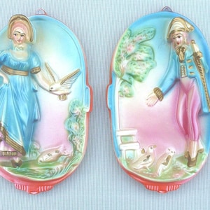 May include: Two vintage wall plaques with a colorful painted design. One plaque features a woman in a blue dress with a white bird, and the other features a man in a red and blue suit with a white bird.