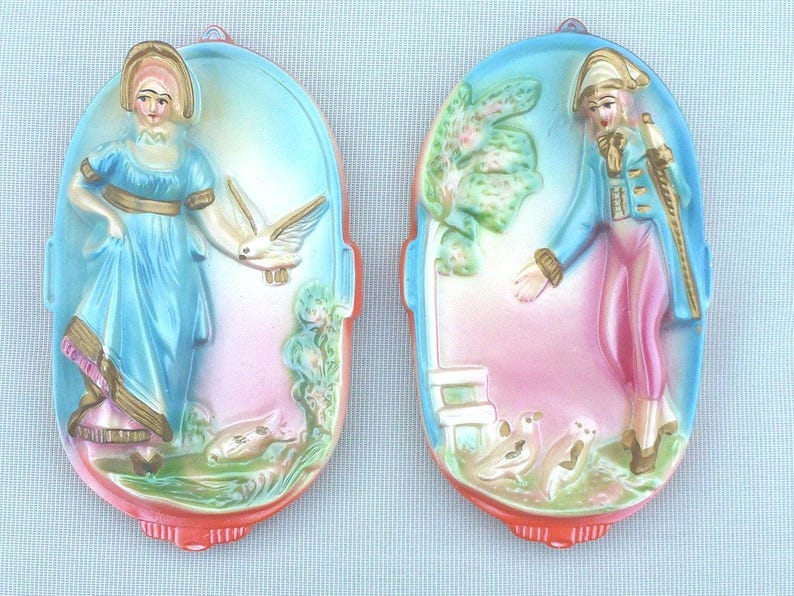 May include: Two vintage wall plaques with a colorful painted design. One plaque features a woman in a blue dress with a white bird, and the other features a man in a red and blue suit with a white bird.