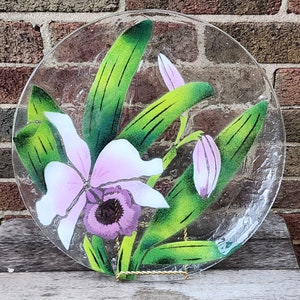 May include: A clear glass plate with a painted design of pink and white orchids with green leaves. The plate is on a wooden surface.