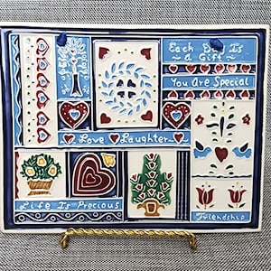May include: A white ceramic tile with a blue border and a patchwork design. The tile features various images, including hearts, flowers, birds, and trees. The tile also includes the text "Each Day Is a Gift", "You Are Special", "Love Laughter", "Life Is Precious", and "Friendship".