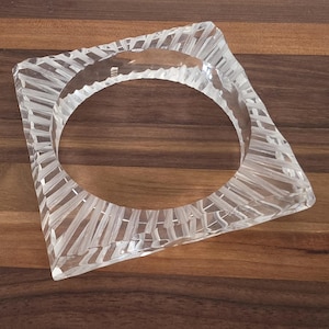 Puede incluir: Brazalete cuadrado transparente con un dise&ntilde;o lineal texturizado. El brazalete tiene una gran abertura ovalada en el centro y est&aacute; hecho de un material transparente. El brazalete se muestra sobre una superficie de madera marr&oacute;n.