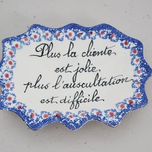May include: A white ceramic plaque with a scalloped edge, featuring a blue and red floral border. The plaque has handwritten black script in French. The text reads: "Plus la cliente est jolie, plus l'auscultation est difficile."