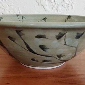 May include: A large, green ceramic bowl with a white glaze and black painted branches and leaves. The bowl is sitting on a wooden surface.