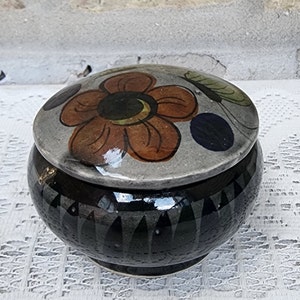 May include: A small, round ceramic trinket box with a lid. The lid is decorated with a hand-painted orange flower with a black center and green leaves. The box is black with a gray rim and a pattern of black triangles.
