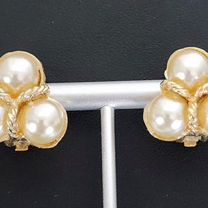 May include: A pair of clip-on earrings featuring three faux pearls in a cluster design. The pearls are cream-colored and set in a gold-tone metal frame with a rope-like texture. The earrings are displayed on a silver stand against a black background.