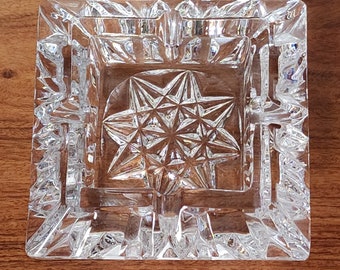 May include: A clear glass ashtray with a starburst design. The ashtray is square and has a raised edge.