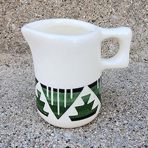 May include: A white ceramic creamer with a green and black geometric pattern. The creamer has a handle and a spout.