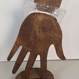 Puede incluir: Un soporte de exhibici&oacute;n de madera de color marr&oacute;n oscuro con forma de mano y base circular, que sostiene un objeto cuadrado de vidrio transparente y texturizado con un patr&oacute;n acanalado en la superficie.
