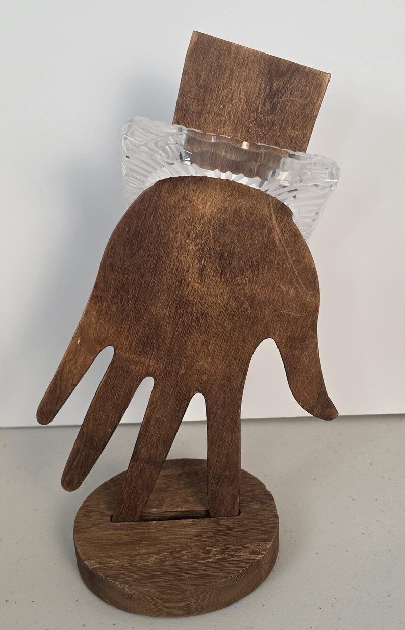 Puede incluir: Un soporte de exhibici&oacute;n de madera de color marr&oacute;n oscuro con forma de mano y base circular, que sostiene un objeto cuadrado de vidrio transparente y texturizado con un patr&oacute;n acanalado en la superficie.