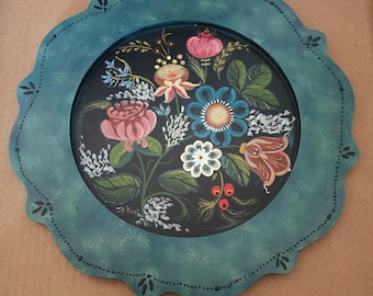 Op de afbeelding: Een handgeschilderde, ronde decoratieve plaat met een geschulpte teal rand. Het midden heeft een bloemenontwerp met roze, blauwe en witte bloemen op een zwarte achtergrond. De plaat is in folk art stijl.