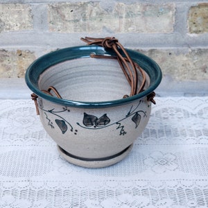 May include: A white ceramic hanging planter with a green rim and brown leather straps. The planter has a floral design with green leaves and brown accents.