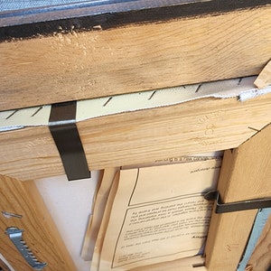 May include: A wooden frame with a canvas inside. The frame is brown and has a black painted edge. The canvas is white and has a piece of paper on it with text that says "This painting is a raw canvas, original art".