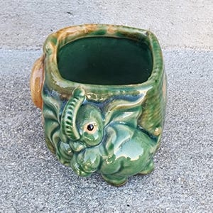 May include: A green ceramic elephant planter with a square opening. The elephant's trunk and face are detailed, with a small eye visible. The planter has a dark green interior and a textured exterior.