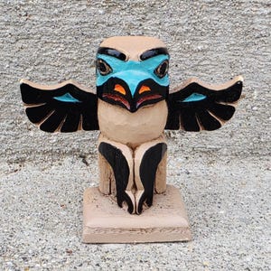 May include: A carved wooden totem figure featuring a stylized bird with outstretched wings. The figure is painted in light tan, black, bright turquoise, and orange accents, standing on a square base with visible wood grain texture throughout.