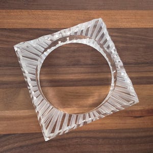Puede incluir: Brazalete cuadrado transparente con una abertura circular. El brazalete est&aacute; hecho de un material transparente con un dise&ntilde;o lineal texturizado. El brazalete se muestra sobre una superficie de madera con rayas marrones claras y oscuras alternas.