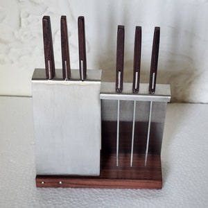 May include: A stainless steel knife block with a wooden base. The block holds six knives with dark wood handles. The knives are arranged in two rows, with three knives in each row.