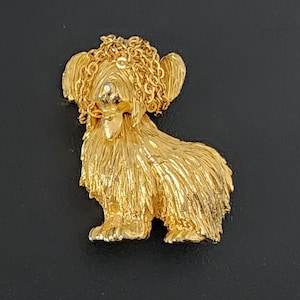 May include: A gold-colored dog brooch with a textured coat and chain-like details around the head and ears. The dog is in a seated position, with a detailed face and a small, round accent on the nose.