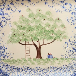 May include: A ceramic plate with a hand-painted design. The central image features a tree with green leaves and red fruit, a ladder, and a basket of fruit. The border has a blue and white speckled pattern.