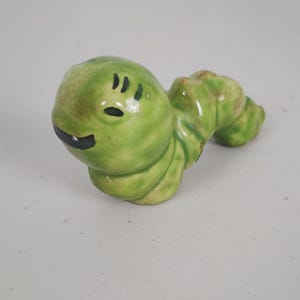 May include: A ceramic caterpillar figurine in shades of green. The caterpillar has a smiling face with black eyes and a mouth. The body is segmented and curved. A whimsical decorative item.