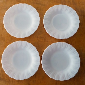 May include: Four white ceramic bowls with a scalloped edge. The bowls are arranged in a square pattern on a wooden surface.