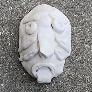 May include: A white, abstract mask with exaggerated features. The mask has large, circular eyes, a prominent nose, and a wide, open mouth with a rectangular shape inside. The surface appears textured, and the mask is likely made of ceramic or a similar material.