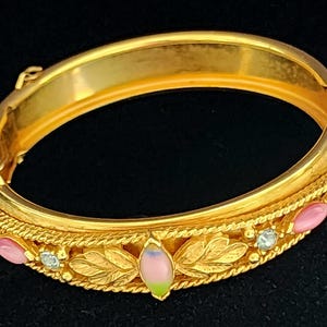 May include: A gold-colored hinged bangle bracelet. The bracelet features a decorative band with pink and blue-green oval-shaped stones, clear round stones, and leaf designs. The bracelet is set against a black background.