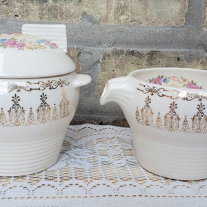 May include: Cream-colored ceramic serving dish and creamer set. The dish has a lid with floral accents and gold trim. The creamer has a spout and handle, also with floral accents and gold trim. Both pieces have a ribbed design.