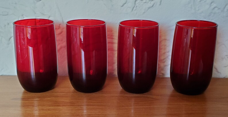 May include: Four red glass tumblers with a dark red base. The glasses are clear and have a smooth, rounded shape.
