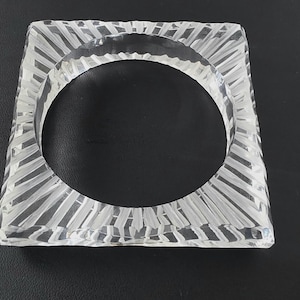 Puede incluir: Un brazalete cuadrado transparente. El brazalete tiene una superficie texturizada con un patr&oacute;n de l&iacute;neas verticales que irradian desde el centro. El brazalete est&aacute; hecho de un material transparente, permitiendo el paso de la luz.