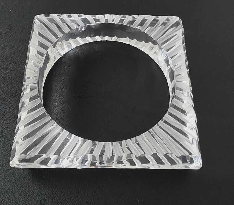 Puede incluir: Un brazalete cuadrado transparente. El brazalete tiene una superficie texturizada con un patr&oacute;n de l&iacute;neas verticales que irradian desde el centro. El brazalete est&aacute; hecho de un material transparente, permitiendo el paso de la luz.