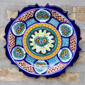 May include: A decorative ceramic plate with a vibrant floral design. The plate features a dark blue scalloped edge and a central pattern of yellow, green, and blue flowers. The plate's design includes a mix of geometric and floral elements.